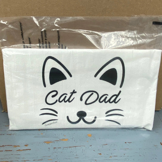White cotton tea towel with 'Cat Dad' printed in black text above a black silhouette of a cat face, placed on a wooden surface with a blurred background.