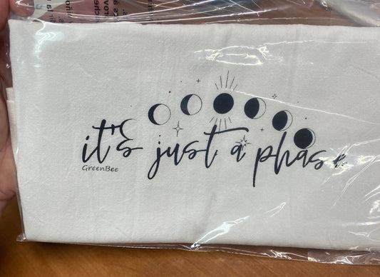 A white cotton kitchen towel with black text and illustrations of moons in different phases. The text reads 'it's just a phase' and there is a small 'Green Bee' logo at the bottom.