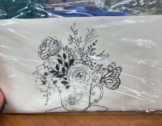 A white cotton kitchen towel with a hand-printed black floral tea cup design, packaged in plastic.