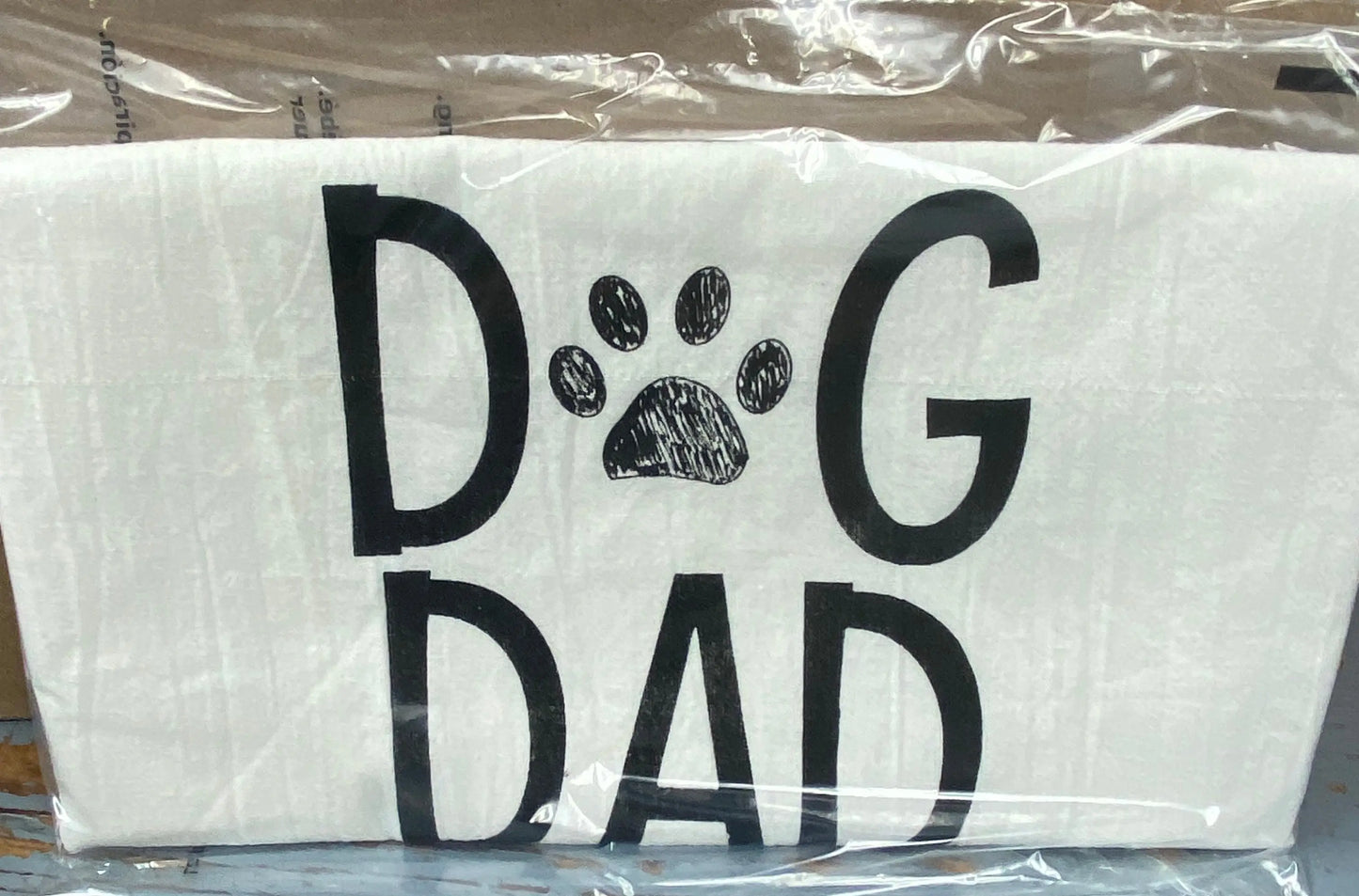 A white cotton tea towel with the words 'DOG DAD' printed in black, with a paw print replacing the letter 'O'.