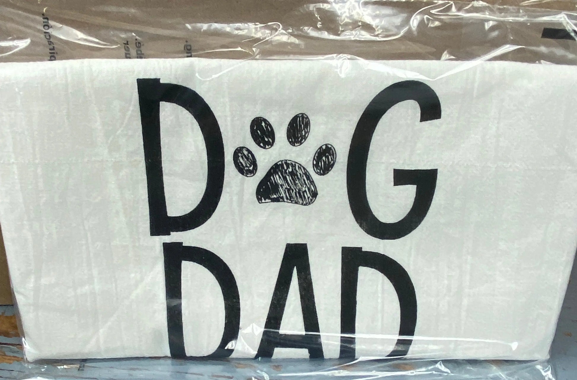 A white cotton tea towel with the words 'DOG DAD' printed in black, with a paw print replacing the letter 'O'.